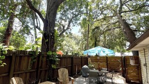 A wooden deck with outdoor furniture, a covered table, and a large tree providing shade. - The Sand Dollar – Charming Bungalow in Gulfport, Minutes from Downtown St. Pete (Gulfport)