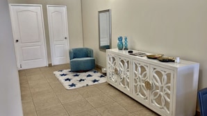 Un corridoio con due porte bianche, una poltrona blu, una consolle bianca con pannelli decorativi, uno specchio e un tappeto a motivi stellari.