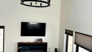 En moderne stue med et stort fladskærms-tv, en grå sofa, en lysekrone og et vindue med skodder.
