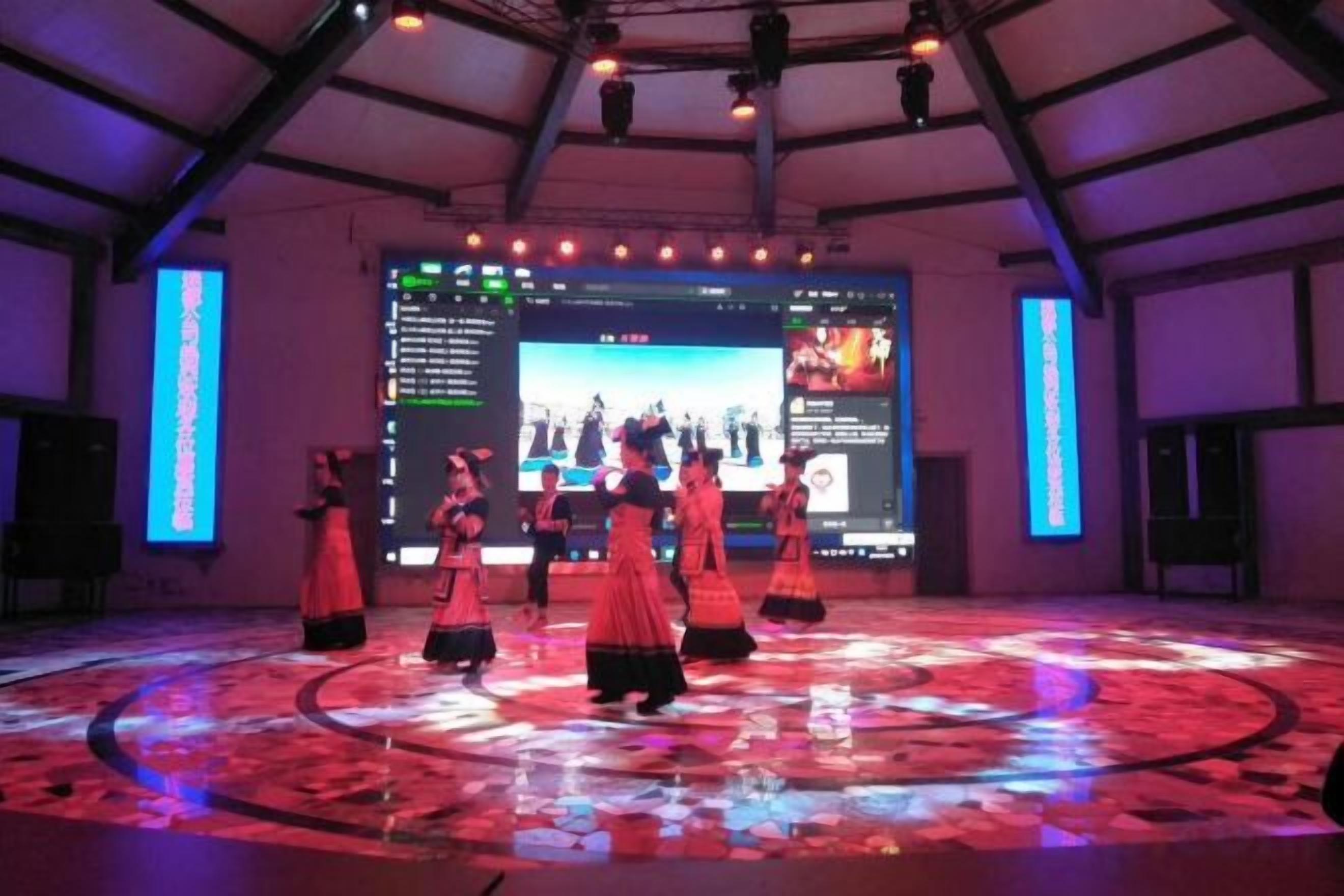 A stage with performers in traditional costumes, a large screen displaying text and images, and a circular patterned floor.