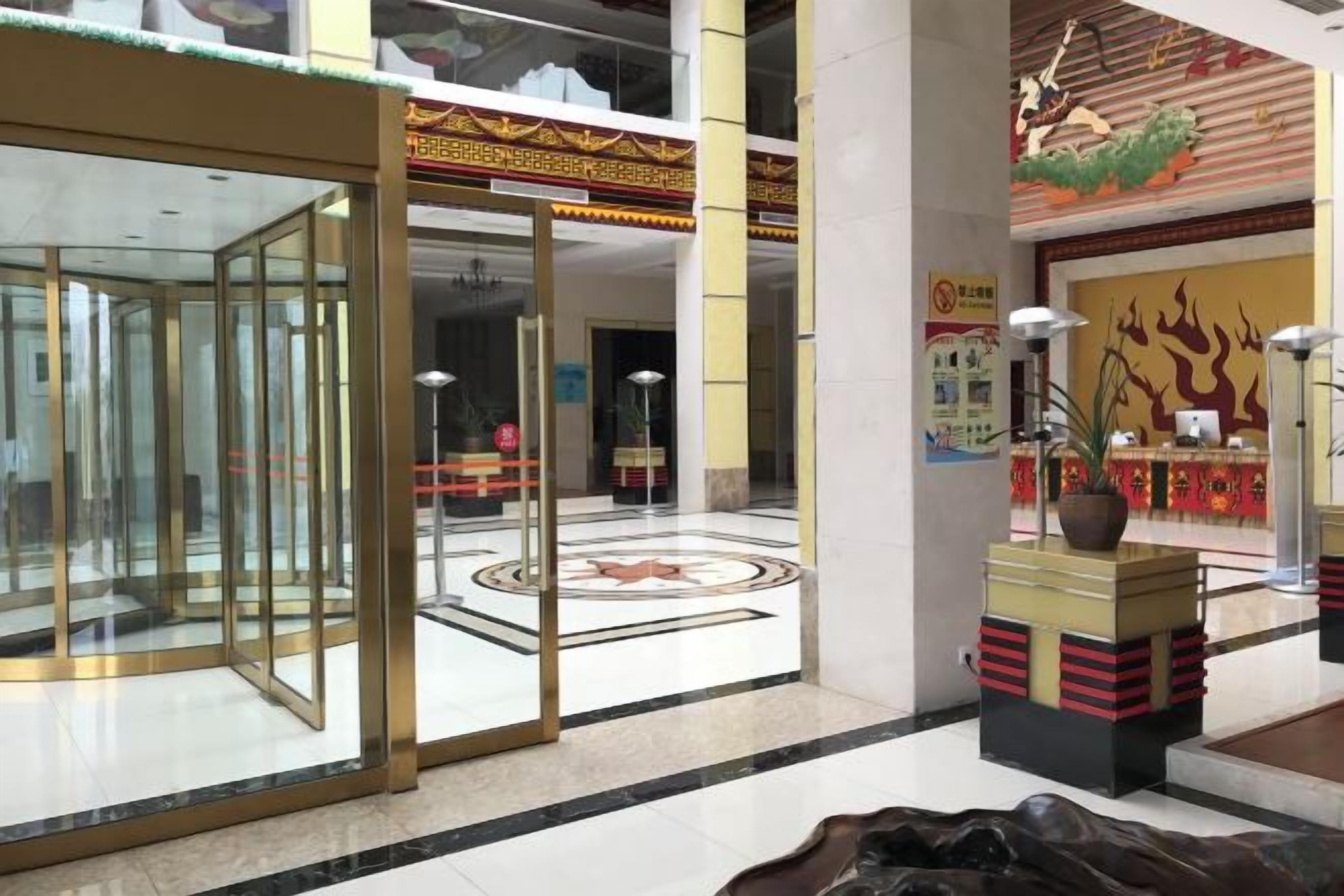 A hotel lobby with a glass partition, a reception desk, a decorative plant, and a traditional mural on the wall.
