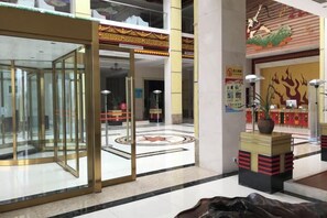 Un vestíbulo de hotel con una partición de vidrio, una recepción, una planta decorativa y un mural tradicional en la pared.