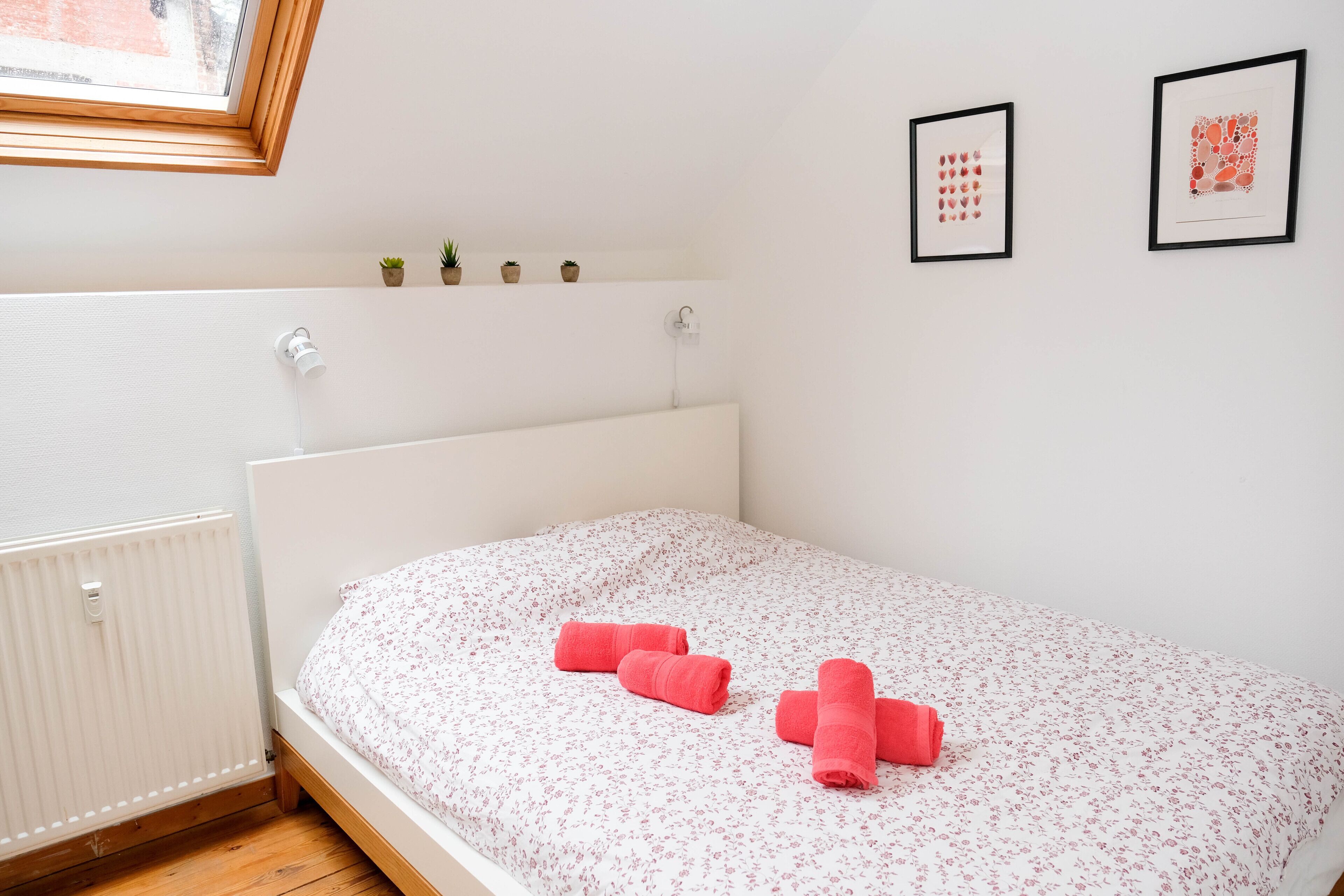 Une chambre avec un lit blanc, une literie à motifs floraux et deux tableaux encadrés accrochés au mur.