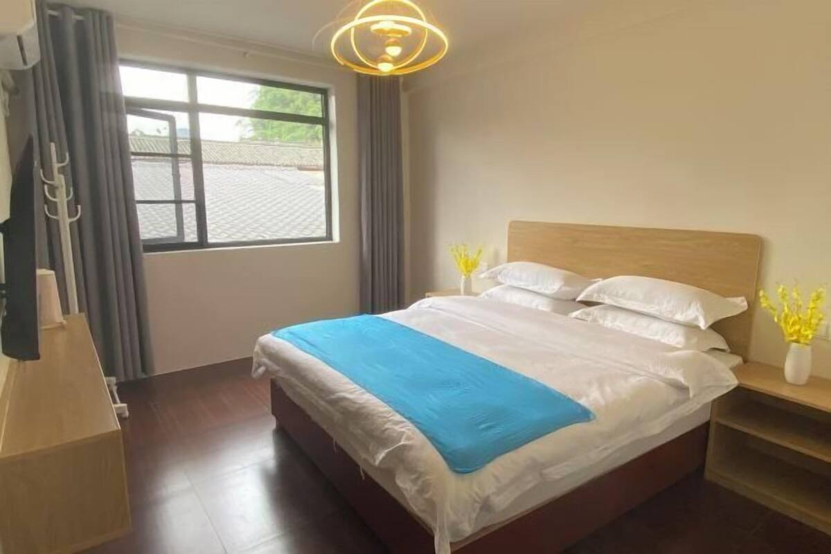 Une chambre d’hôtel avec un grand lit, une tête de lit en bois, une table de chevet ornée d’un vase de fleurs et une fenêtre avec des rideaux.