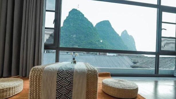 Une pièce dotée d’une grande fenêtre, d’un lit recouvert d’une couvre-lit à motifs, de deux poufs en osier et offrant une vue sur un paysage montagneux.
