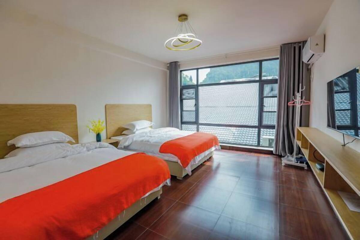 Une chambre d’hôtel avec deux lits, une grande fenêtre, un téléviseur et un banc en bois.