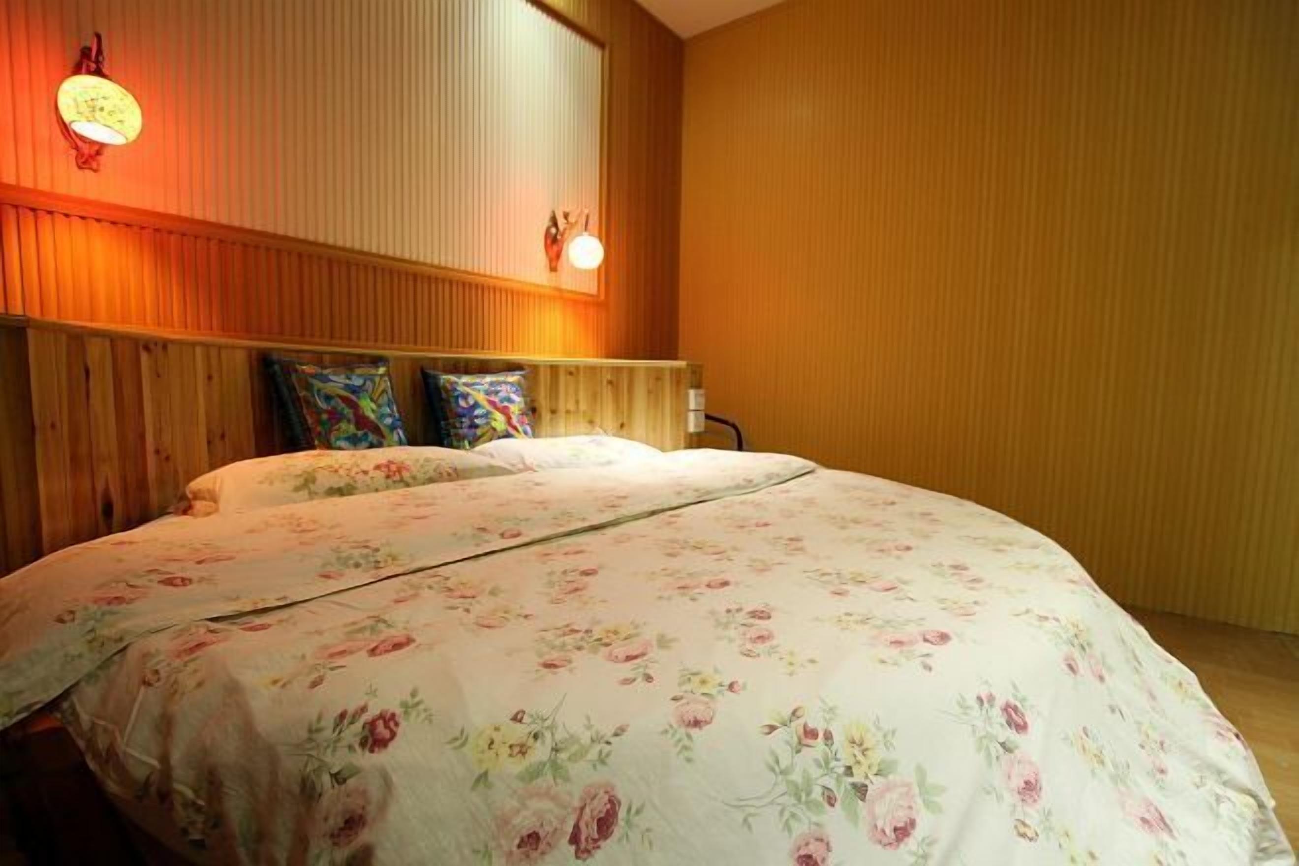 A hotel room with a wooden headboard, floral bedding, and a wall-mounted light fixture.