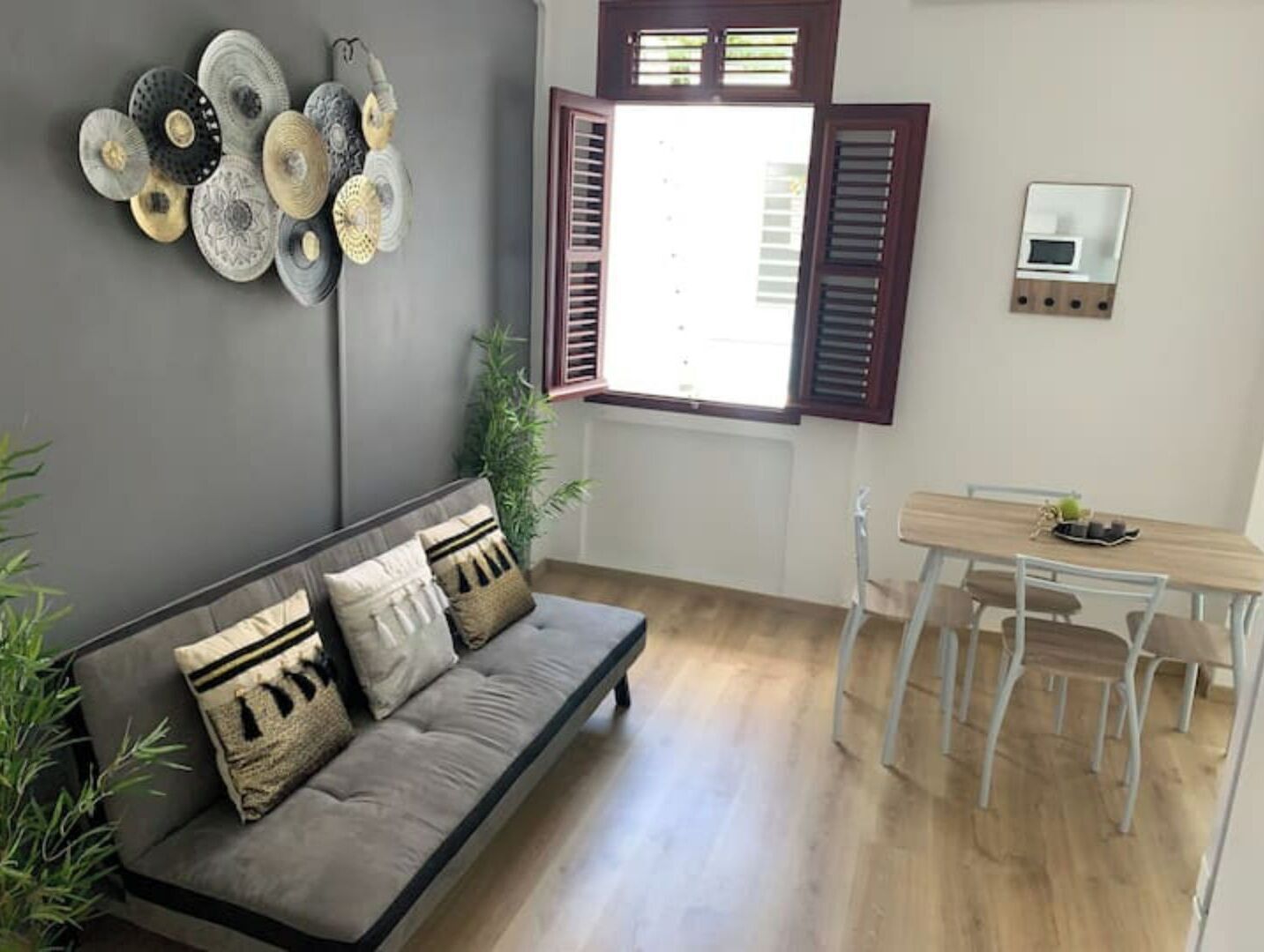 Uma sala de estar com um sofá cinza, uma mesa de jantar com cadeiras, uma janela com persianas e obras de arte nas paredes.