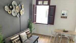 En stue med en grå sofa, et spisebord med stole, et vindue med skodder og billedkunst på væggen.