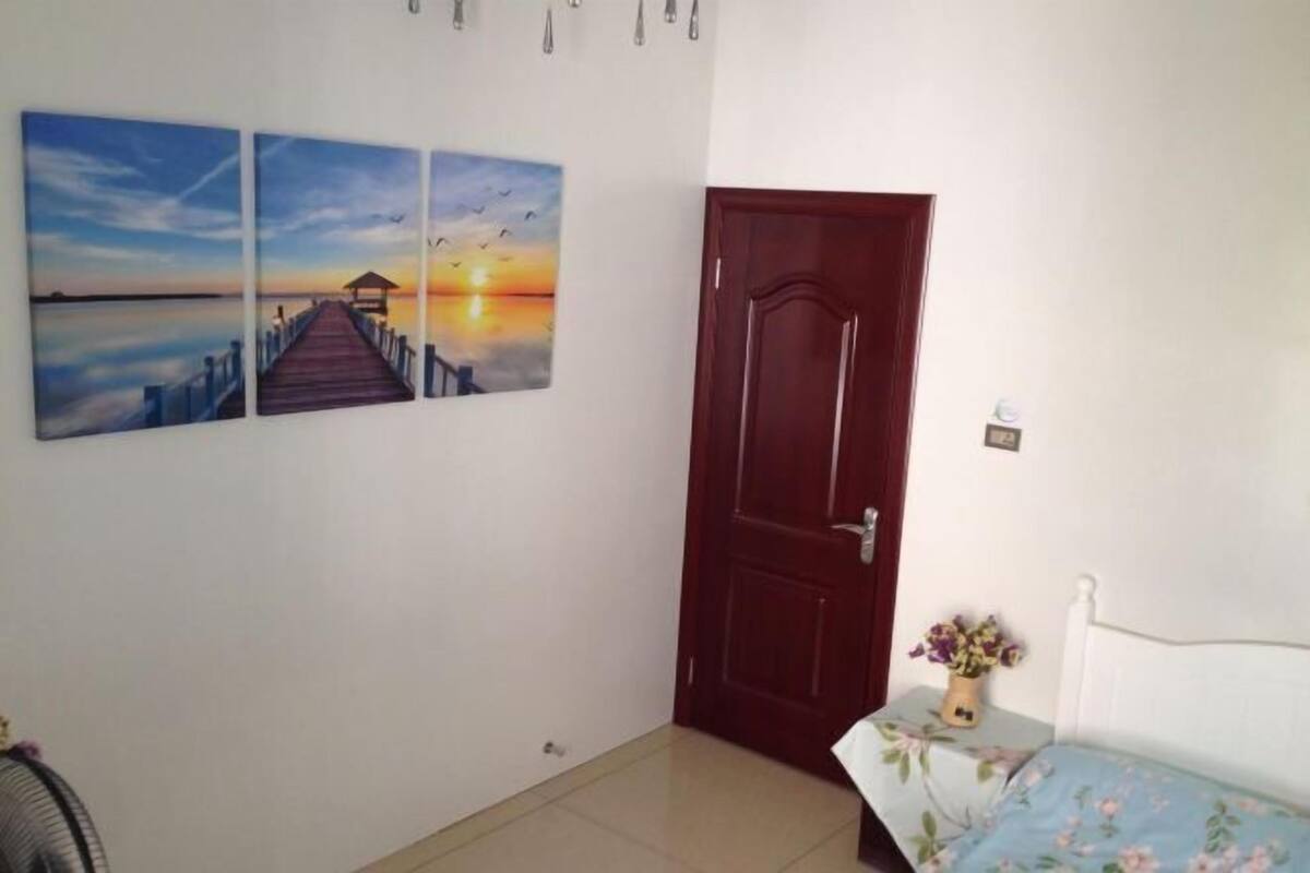 Ein Zimmer mit Holztür, einem Bett mit floralem Überwurf und drei an der Wand hängenden Bildern, die einen Sonnenuntergang über einem Steg zeigen.