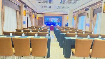 Uma sala de conferências com fileiras de mesas e cadeiras, um palco com fundo azul e uma tela grande.