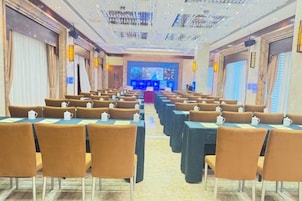Un salón de conferencias con filas de mesas y sillas, un escenario con fondo azul y una pantalla grande.
