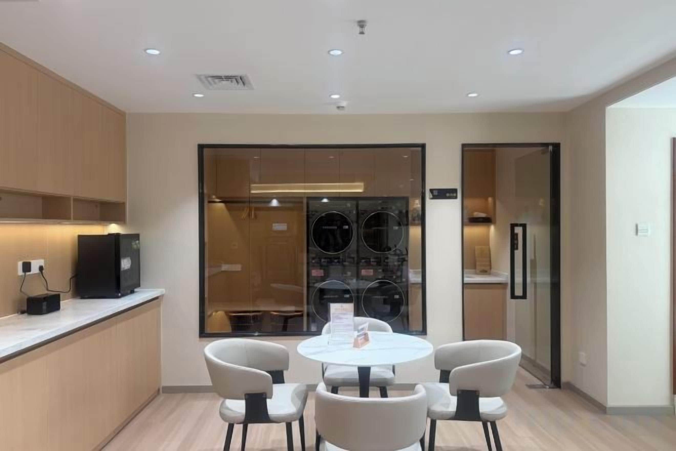 Une chambre d’hôtel moderne avec une table de salle à manger ronde, quatre chaises, un coin cuisine équipé d’un réfrigérateur et une cloison en verre séparant l’espace repas de la cuisine.