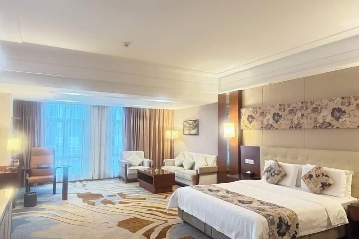 Une chambre d’hôtel avec un grand lit, deux fauteuils, une petite table et une vue sur la ville grâce à de grandes fenêtres.
