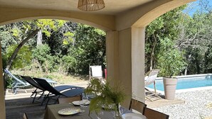 An outdoor dining area with a table set for six, surrounded by chairs, a hanging light fixture, and a view of a pool and trees. - Pleasant house in a quiet area, swimming pool, air conditioning, WIFI, max 12 pers. (Uchaux)