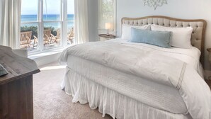 A bedroom with a large bed, a ceiling fan, a view of the ocean through the window, and a decorative mirror on the wall.
