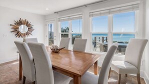 A dining area with a wooden table and white chairs, large windows with a view of the ocean, and a sunburst mirror on the wall.