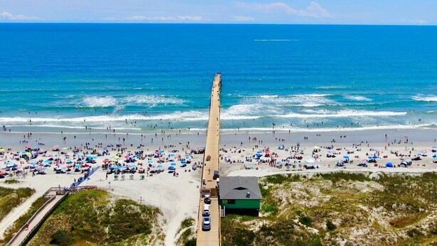 Una spiaggia con un lungo molo che si estende nell'oceano, circondata da persone e ombrelloni.