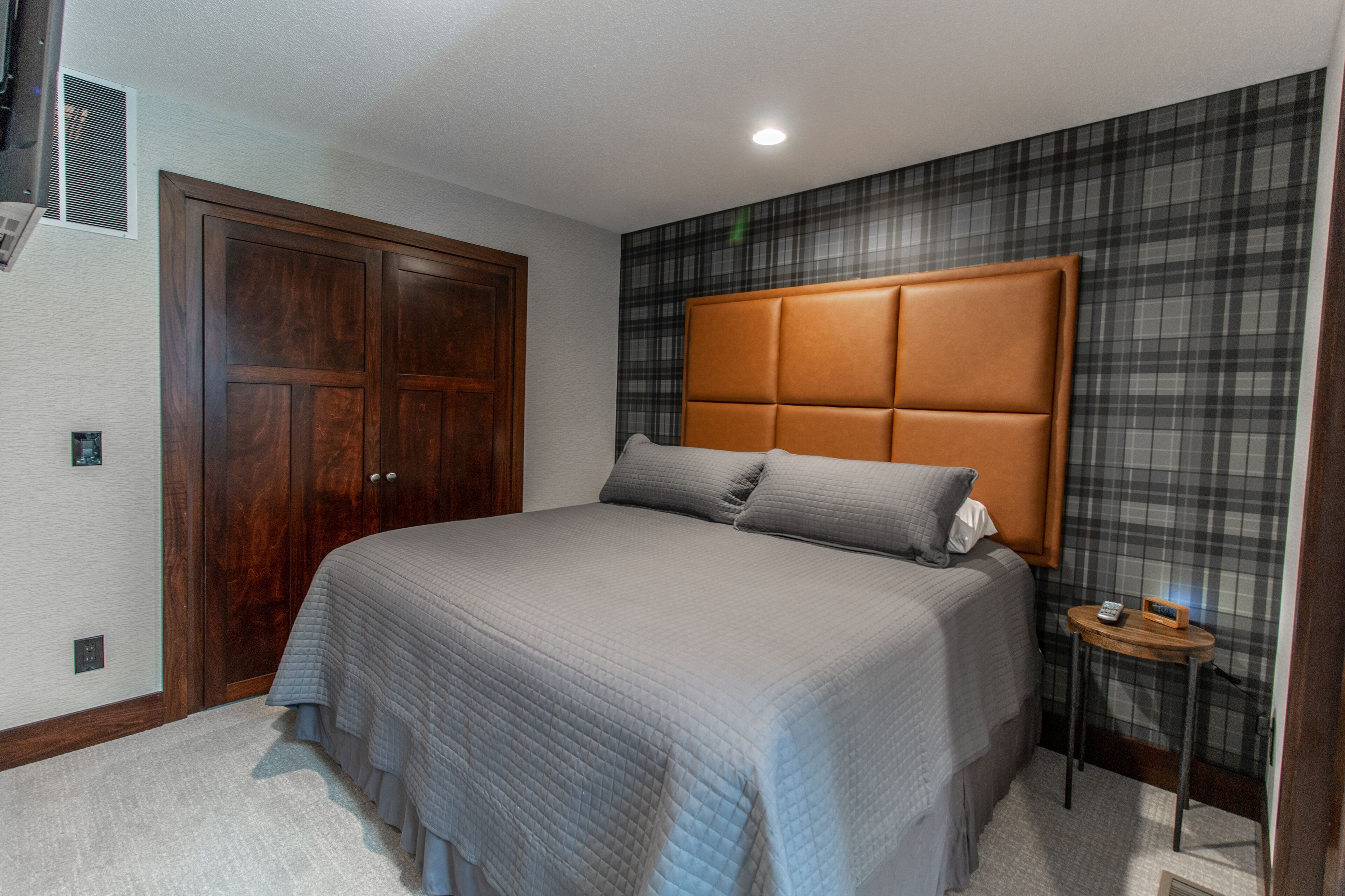 A bedroom with a large bed, a wooden wardrobe, a headboard with a padded section, and a small bedside table with a lamp.