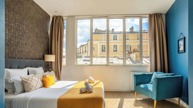 A hotel room with a bed, a blue armchair, a large window with a view of buildings, and a patterned wall.