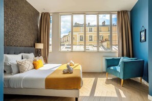 Habitación de hotel con una cama, un sillón azul, un ventanal con vistas a edificios y una pared con diseño.