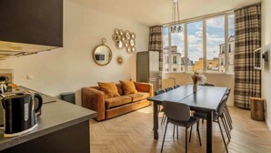 A modern kitchen with a kettle, a sofa, a dining table, and a large window with curtains.