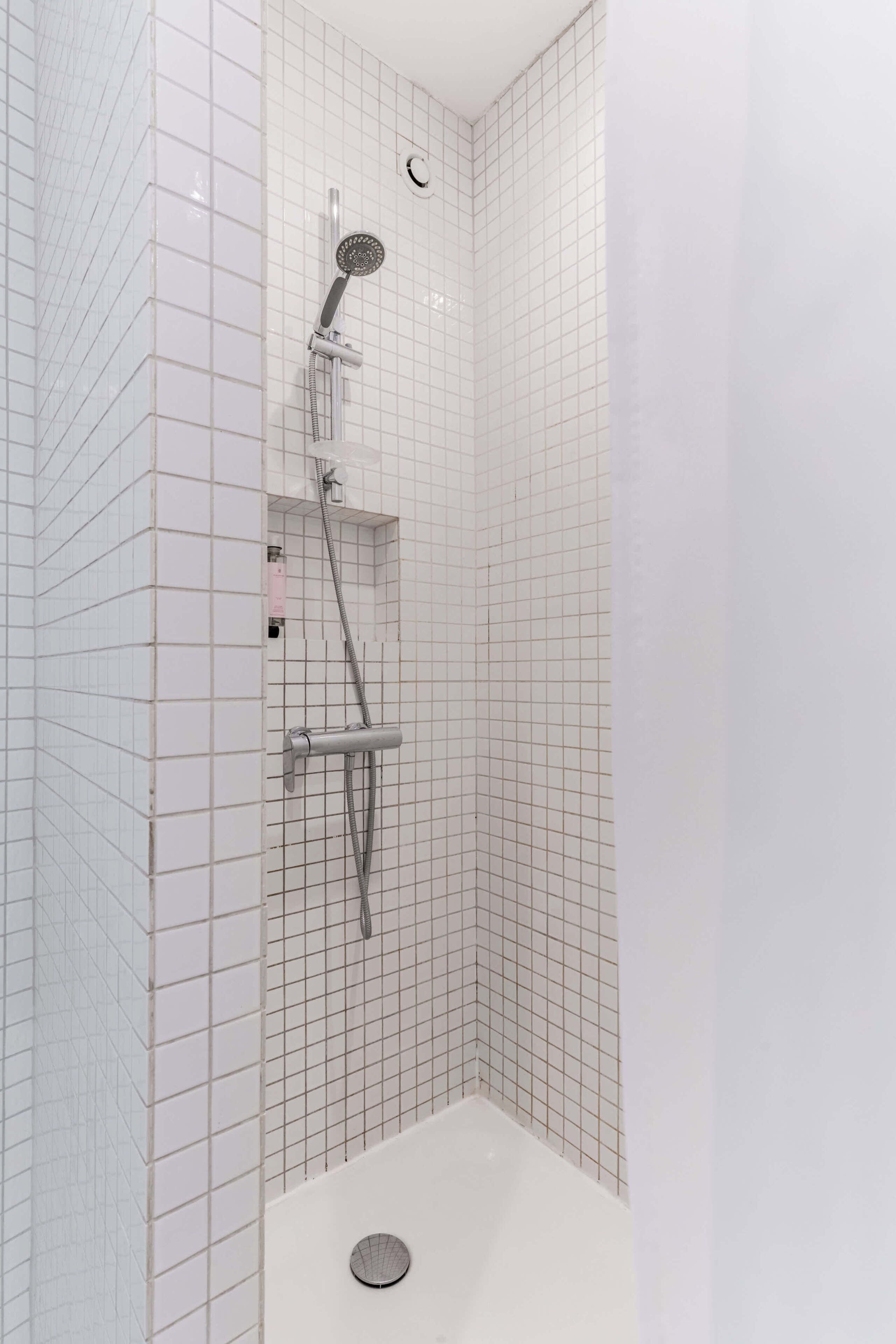 Une douche équipée d’un pommeau de douche à main et d’un robinet mural, des murs carrelés et une baignoire blanche.