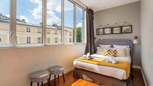 Une chambre avec une grande fenêtre, un lit doté d’une tête de lit, deux tabourets et des tableaux encadrés accrochés au mur.