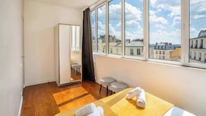 Une chambre d’hôtel avec une grande fenêtre, un lit, une petite table, un miroir et une armoire.