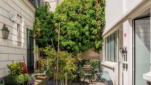Une ruelle étroite en milieu urbain, bordée de bâtiments des deux côtés, un arbre au feuillage dense et une porte avec une enseigne rouge.