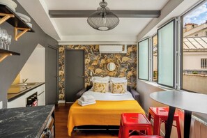 A bedroom with a bed, a window, a red stool, and a kitchen area.