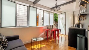 Un espace de vie moderne comprenant un canapé, une table en verre, des tabourets rouges, une poubelle noire et un coin cuisine doté d’un plan de travail et d’étagères noirs.