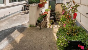 Une ruelle étroite bordée de bâtiments des deux côtés, un petit espace jardin avec des plantes et des fleurs en pot, et une porte visible donnant sur une cour.