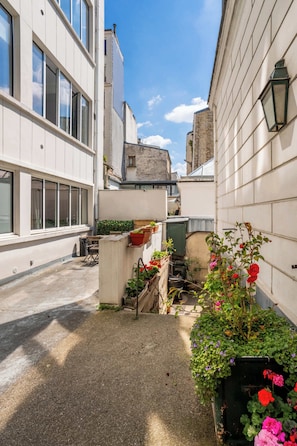 Une ruelle étroite bordée de bâtiments des deux côtés, un petit espace jardin avec des plantes et des fleurs en pot, et une porte visible donnant sur une cour.