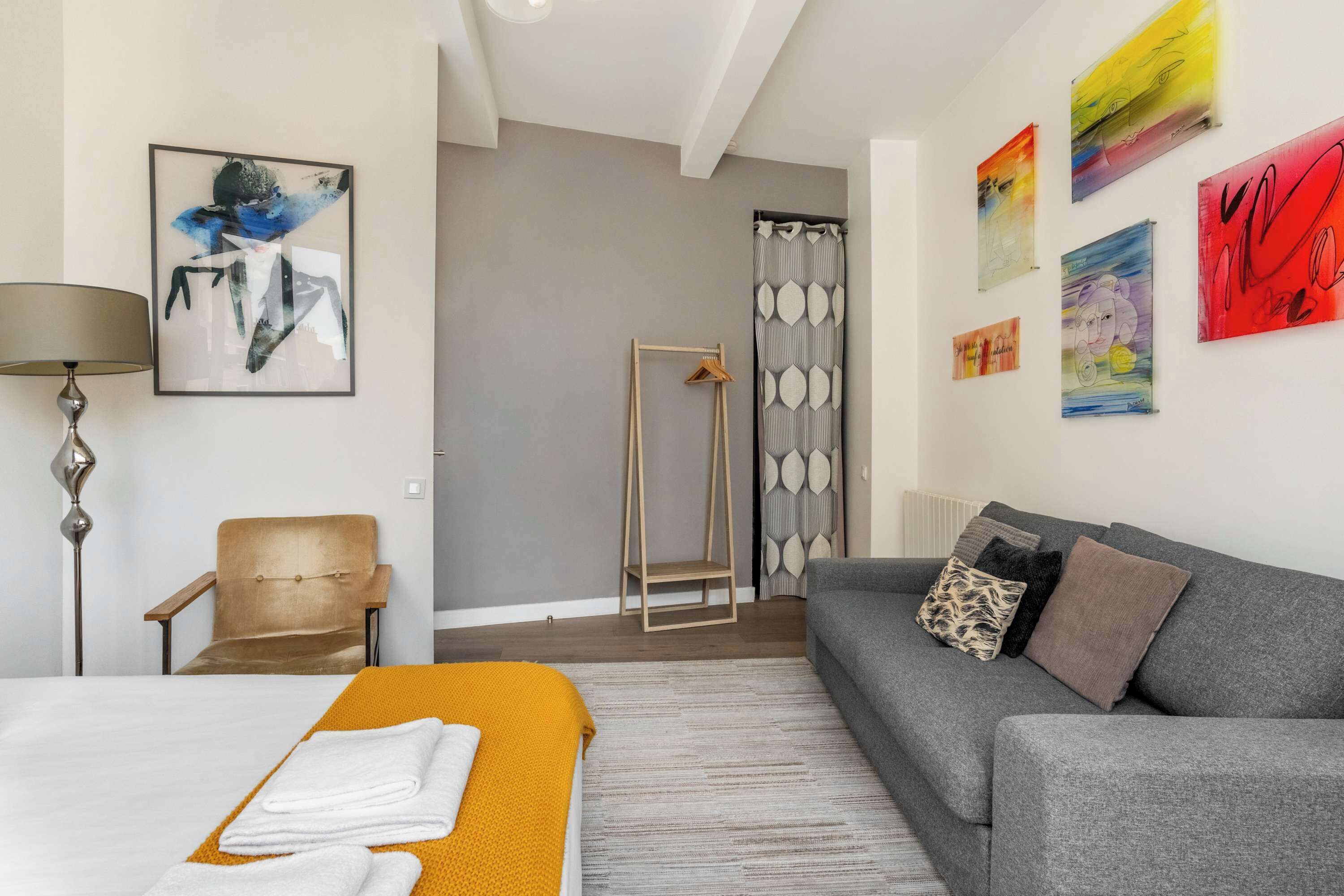 A modern hotel room with a bed, a sofa, a chair, a standing shower, and artwork on the walls.