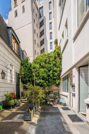A narrow urban alley with buildings on either side, a tree with dense foliage, and a door with a red sign.