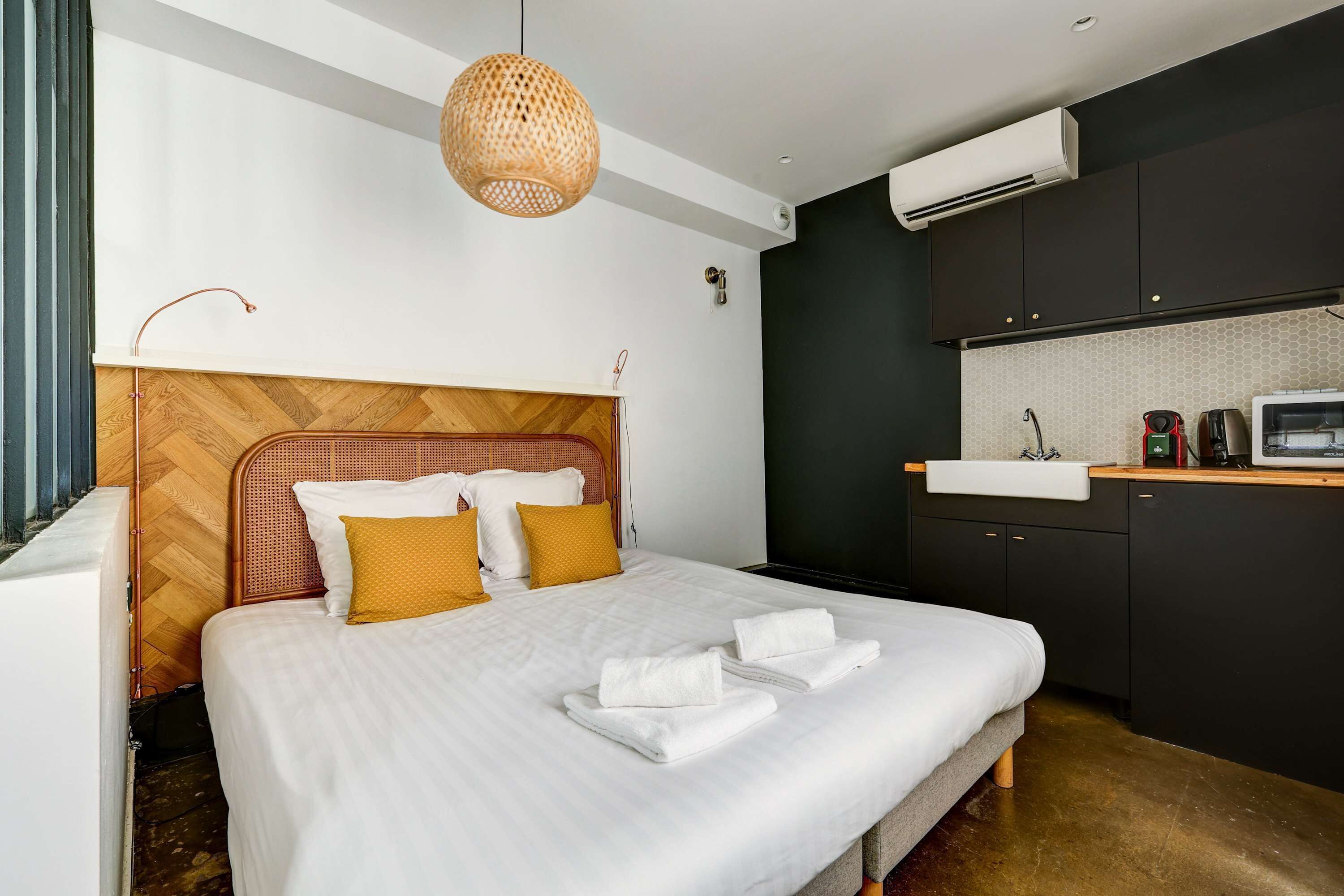 A modern hotel room with a large bed, a wooden headboard, a woven pendant light, a built-in sink, and a flat-screen TV.