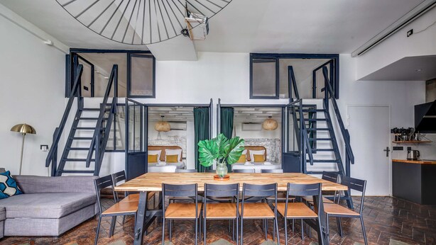 A modern dining area with a wooden table and chairs, a grey sofa, and a staircase leading to an upper level. - Stylish and sunny 7BD with garden for 17 guests! (Paris)