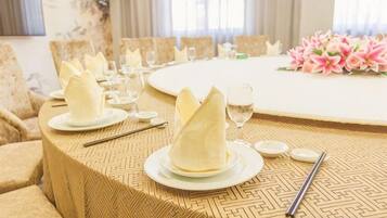 A dining table set with folded napkins, plates, and cutlery, with a floral arrangement in the background.