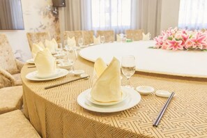 Une table à manger dressée avec des serviettes pliées, des assiettes et des couverts, et un arrangement floral en arrière-plan.