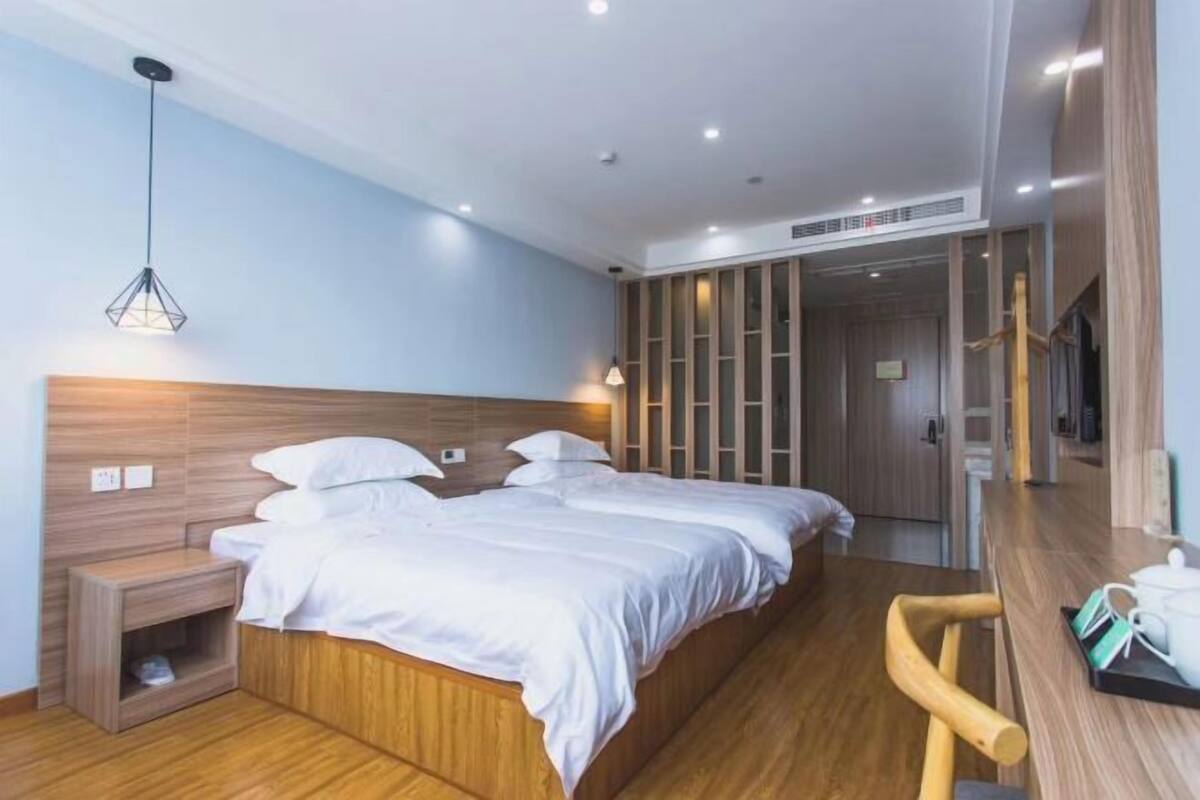 Une chambre d’hôtel moderne avec une tête de lit en bois, deux lits, un bureau en bois, une chaise et un grand miroir.