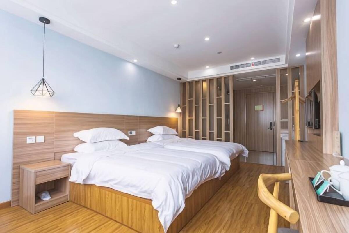 Une chambre d’hôtel avec une tête de lit en bois, deux lits, une table de chevet, une petite étagère et un banc en bois.