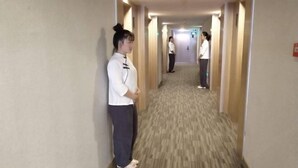 Un couloir d’hôtel avec des revêtements de sol et des murs recouverts de moquette, un détecteur d’incendie sur le côté droit, et une personne se tenant au loin.