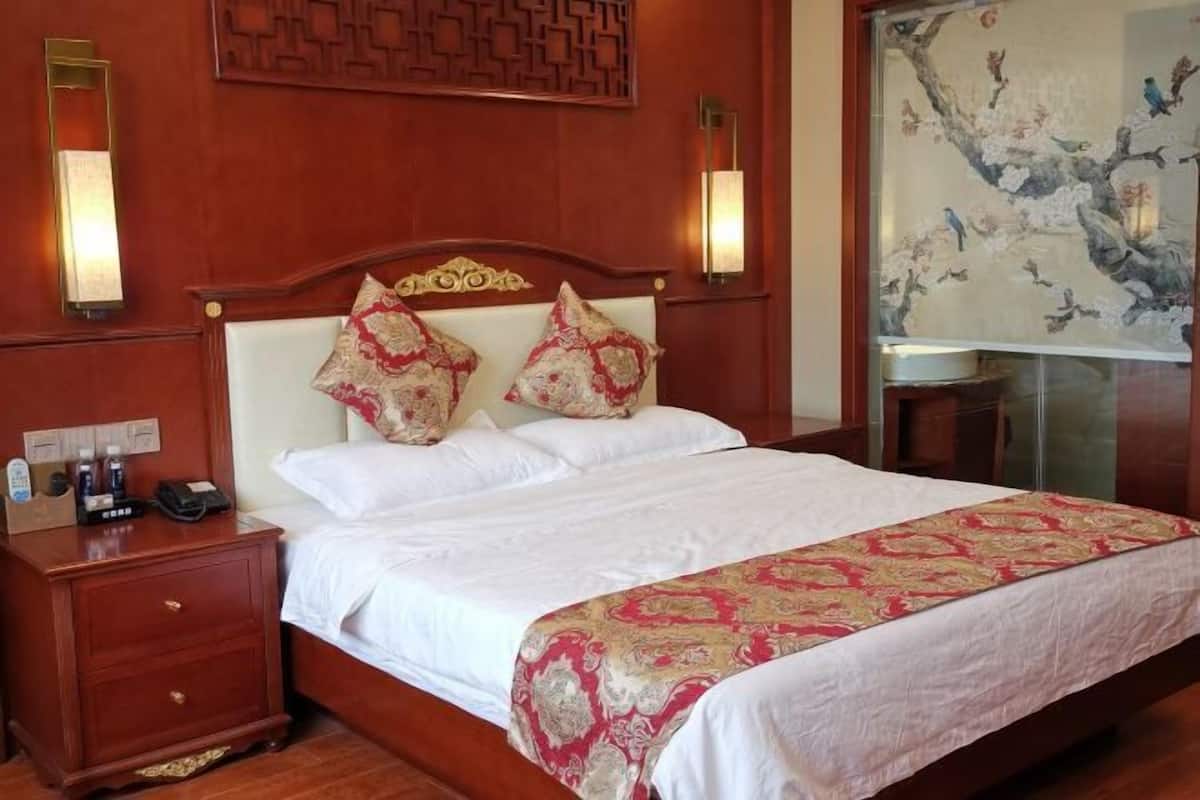 Quarto de hotel com cabeceira de madeira, duas almofadas decorativas, mesa de cabeceira com telefone e garrafas, e luminária fixada na parede.
