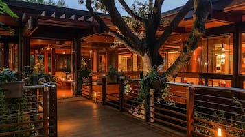 A wooden deck with a glass enclosure, surrounded by trees and plants, leading to a building with large windows and a warm interior glow.