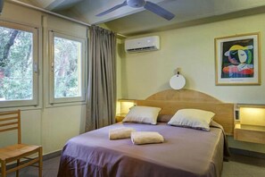 Une chambre d’hôtel comprenant un lit, un ventilateur de plafond, un appareil de climatisation, une fenêtre avec des rideaux, une chaise et un luminaire fixé au mur.