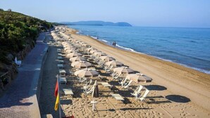 A beach with rows of sun loungers and umbrellas, a paved walkway, and a clear view of the sea and distant hills.