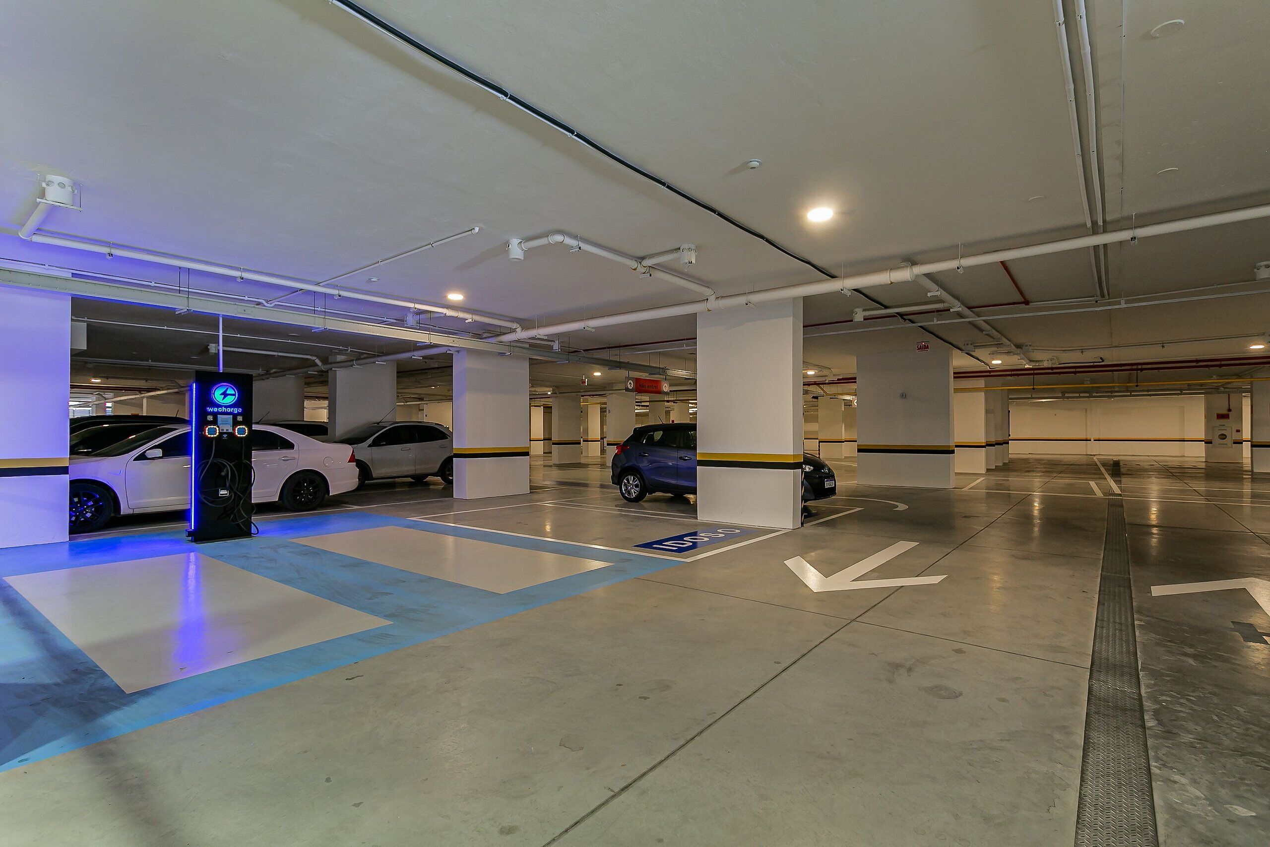 An underground parking garage with clear directional arrows on the floor, a charging station, and several parked cars.