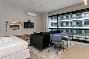 A modern hotel room with a bed, a black sofa, a small table with two chairs, a large window with a view of a building, and a mounted TV. - Pool with Sea View | WI-FI 200MB | Foodhall #TT2 (Florianópolis)