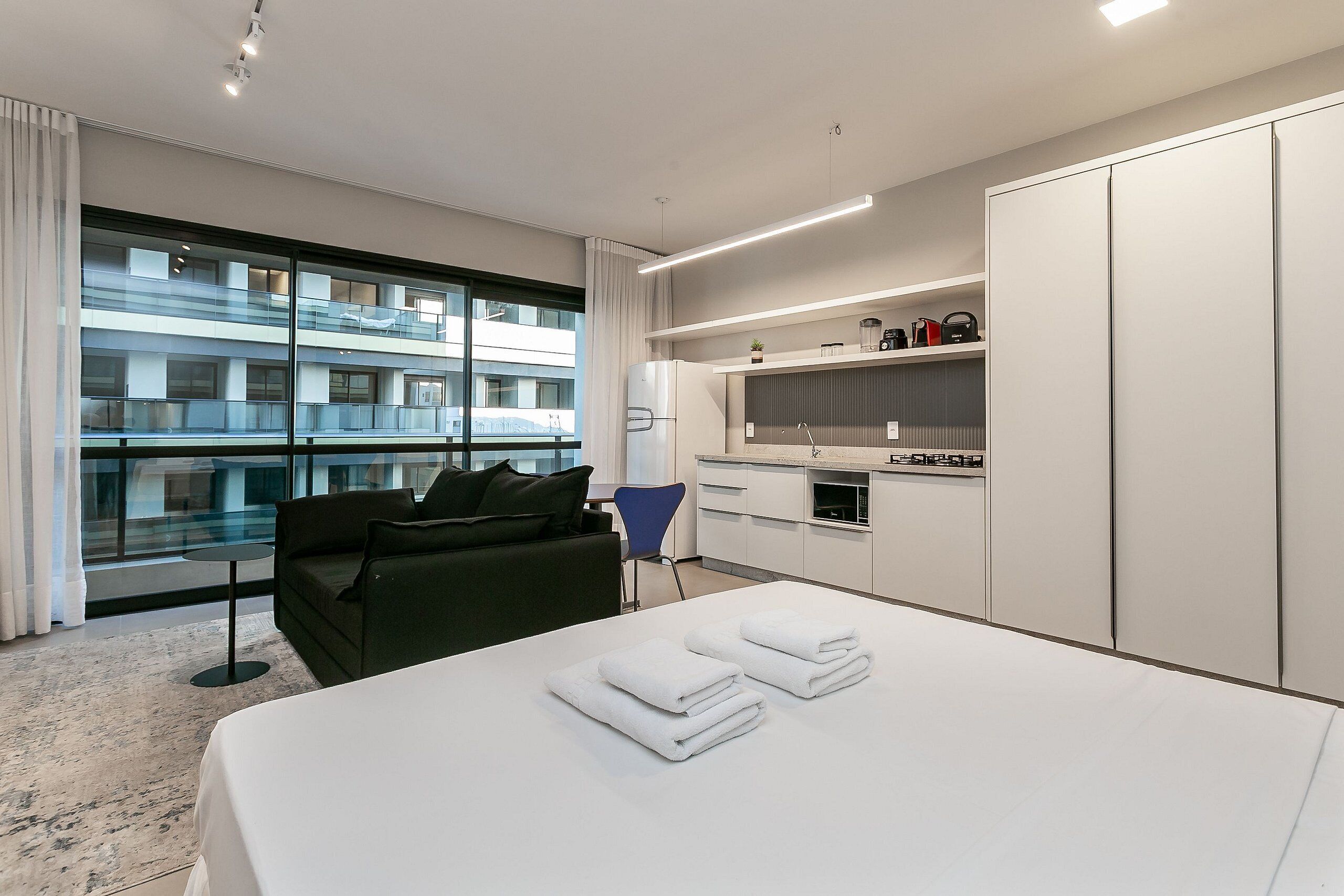A modern hotel room with a large bed, a sofa, a small table, and a kitchen area.